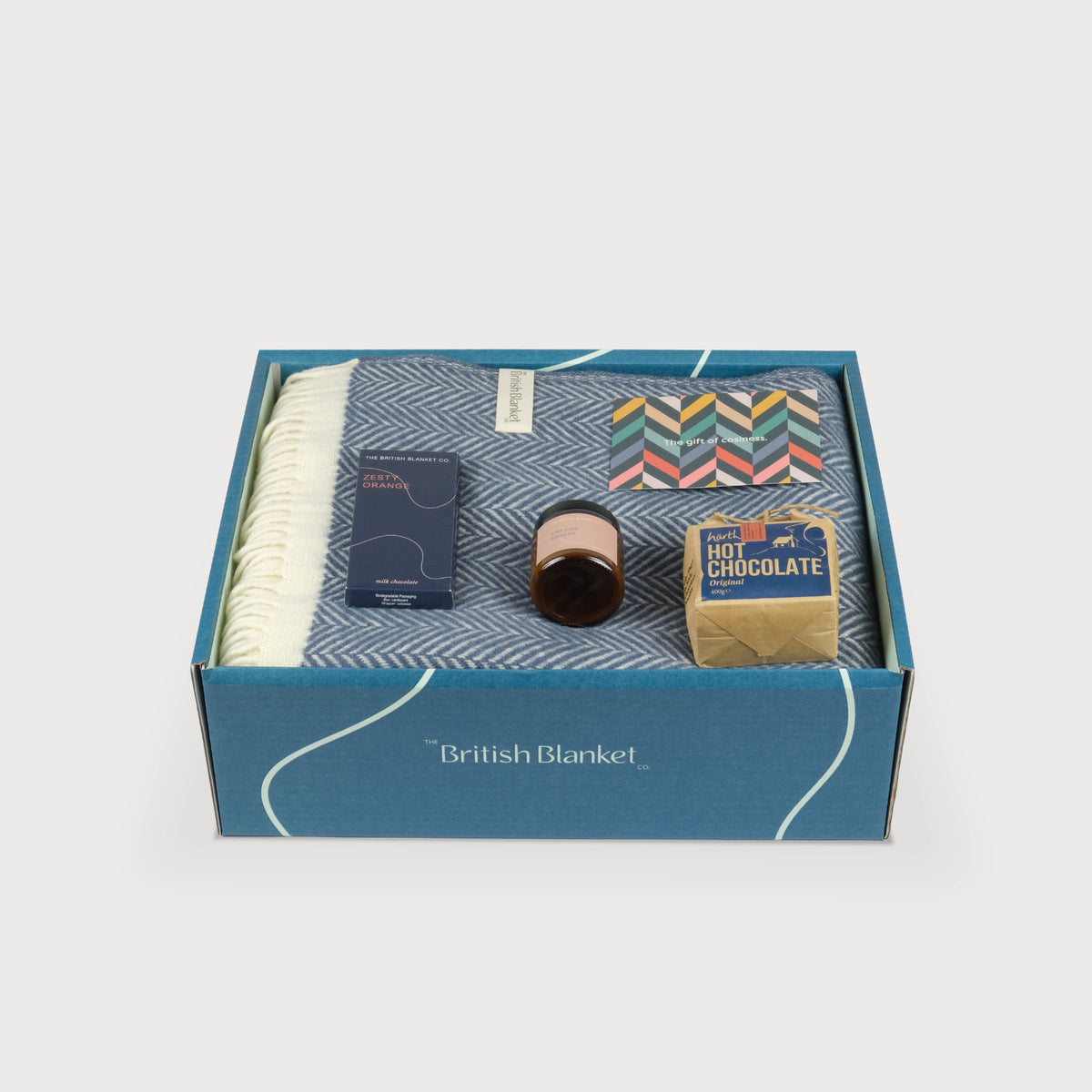 A luxury gift box with a blue herringbone blanket, scented candle, hot chocolate and bar of orange milk chocolate