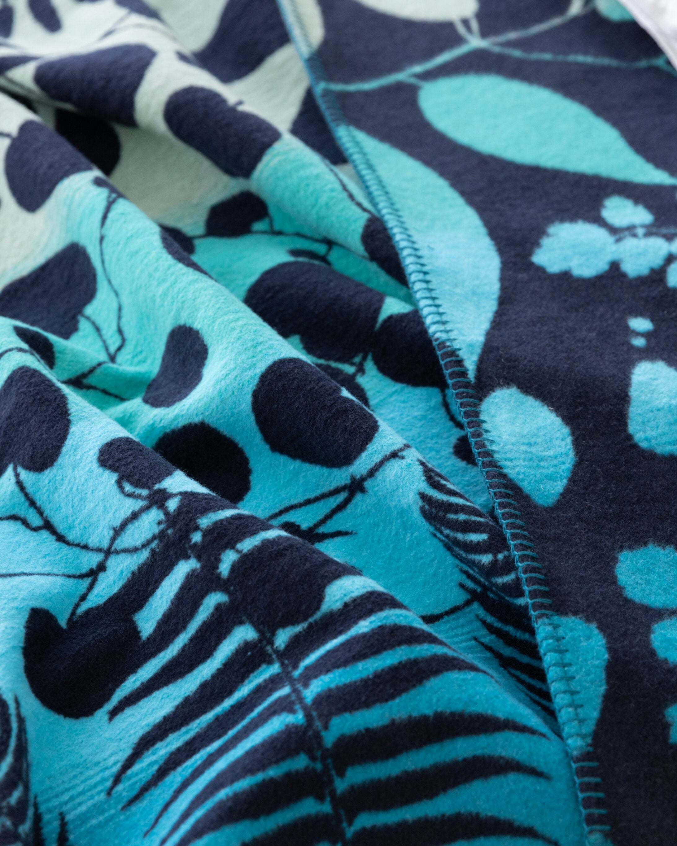 Clarissa Hulse Blanket: Indian Summer in Peacock, featuring teal and navy foliage and dot patterns, with overlock stitching on soft, reversible recycled cotton.