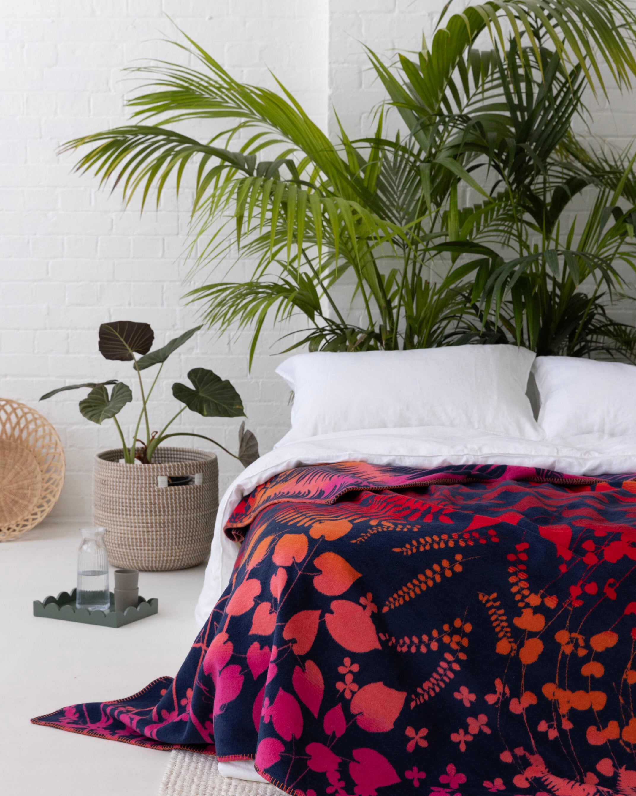 Clarissa Hulse Blanket: Indian Summer in Sunset, a vibrant botanical-patterned throw on a minimalist bed, complemented by lush plants and soft lighting.