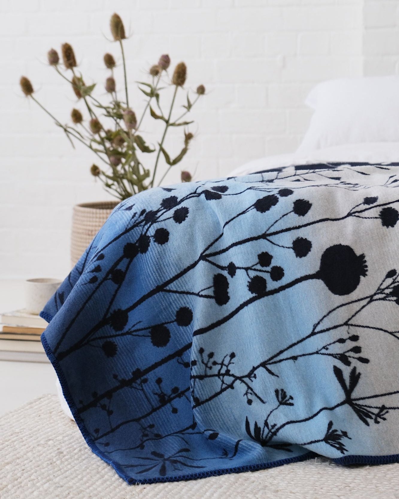 Clarissa Hulse Blanket: Seedheads in Midnight draped over bed, featuring a botanical pattern. White bedding, woven basket with dried flowers, and books complete the cozy scene.