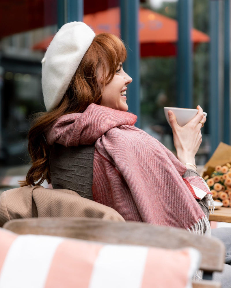 Woman at café wearing Oversized Blanket Scarf: Tabasco Herringbone, wrapped around her shoulders, holding coffee beside a floral bouquet and pastry.