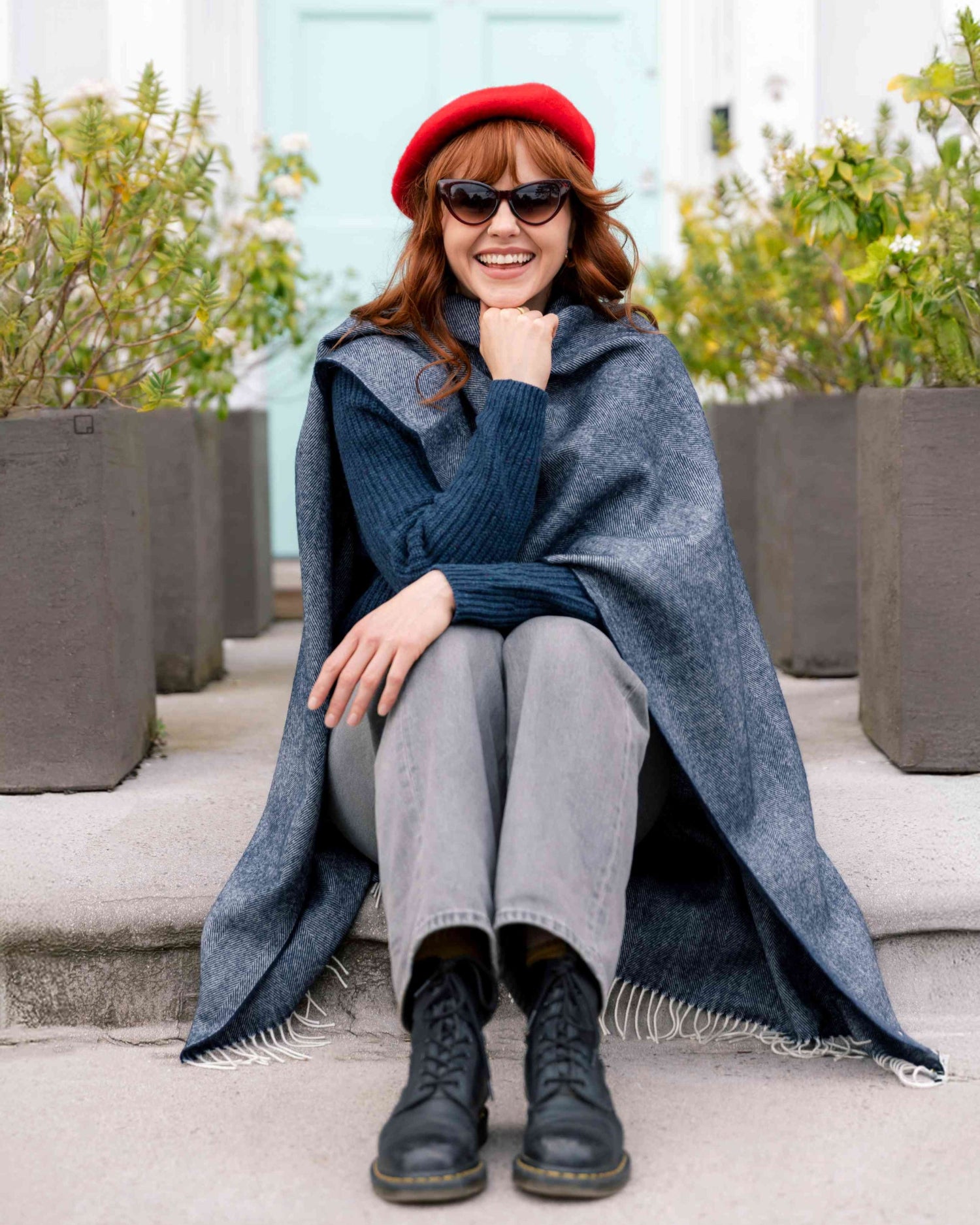 Young woman on steps wearing a Wool Beret Hat: Red, paired with sunglasses, a navy sweater, gray jeans, and combat boots, accentuating her chic, casual style.