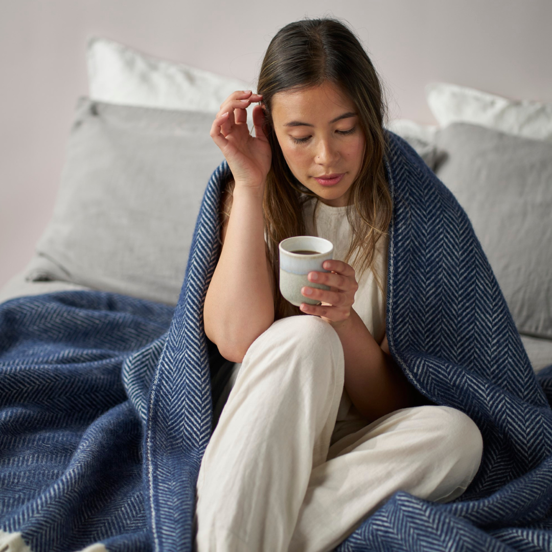 Woman sitting on a couch wrapped in a Clifton Herringbone blanket in Brunel, holding a mug.