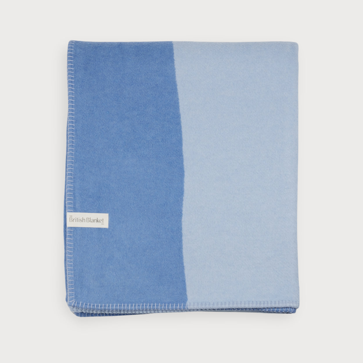 Cloud Cotton Blanket: Tide in Cornflower, folded, showing reversible darker and lighter blue sides with whip-stitch edging and “The British Blanket Co.” label.
