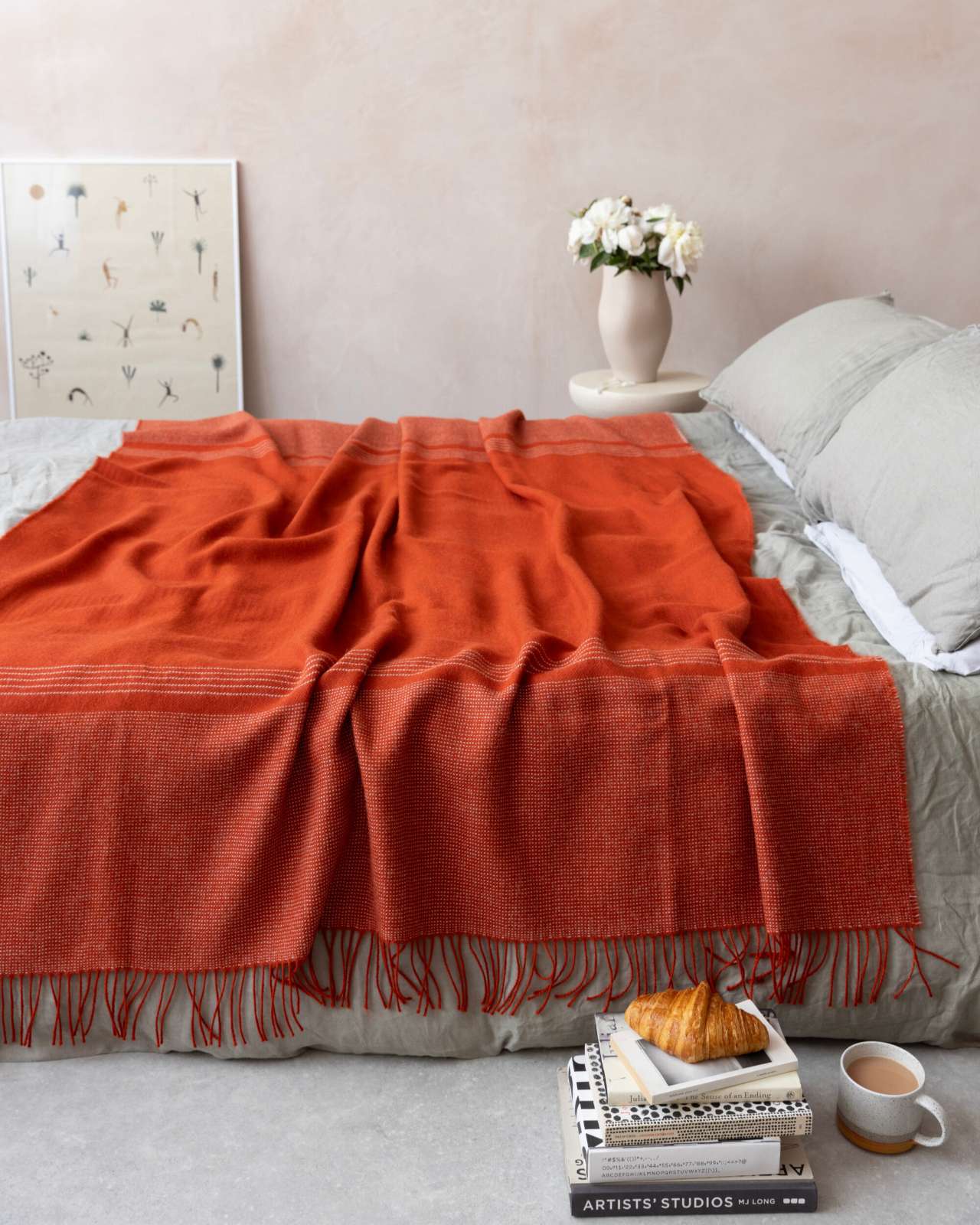 Alpaca Blanket Gift Box: Flame Orange displayed on a bed, featuring a woven throw, art books, and a coffee mug, creating a cozy, modern bedroom scene.