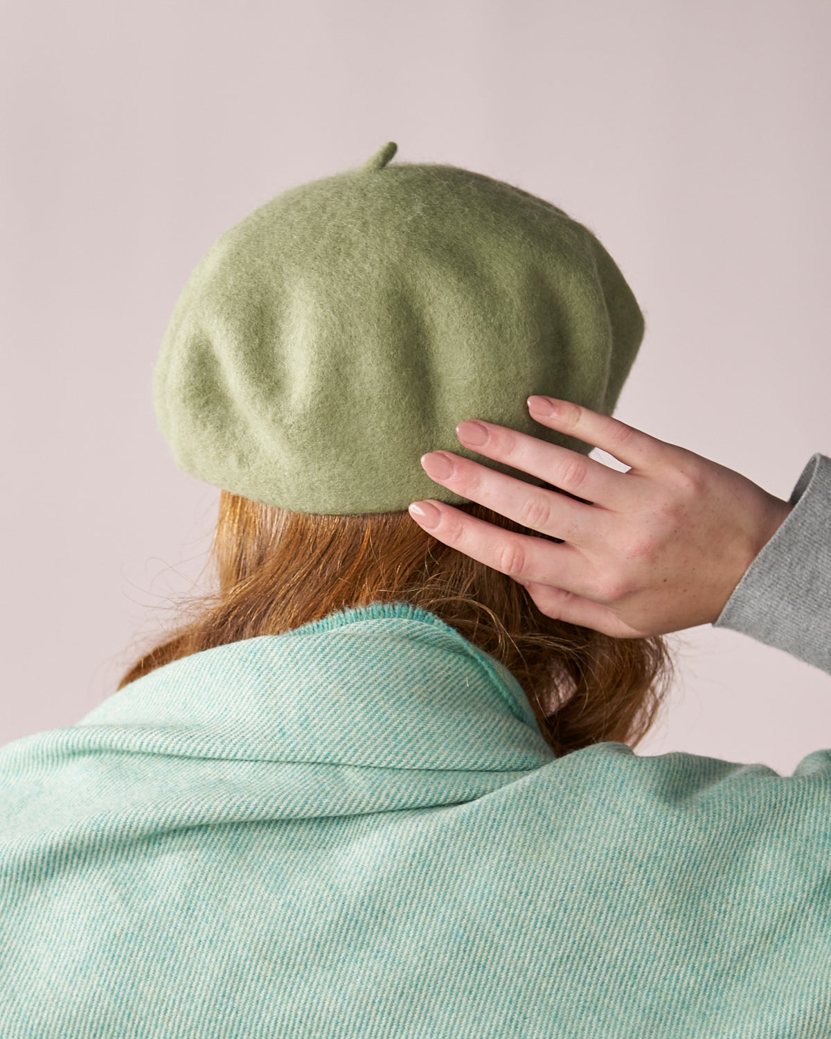 Person wearing a Wool Beret Hat: Light Green, showcasing its thickness and shape. Hand delicately touching the beret, complemented by a textured teal shawl or coat.