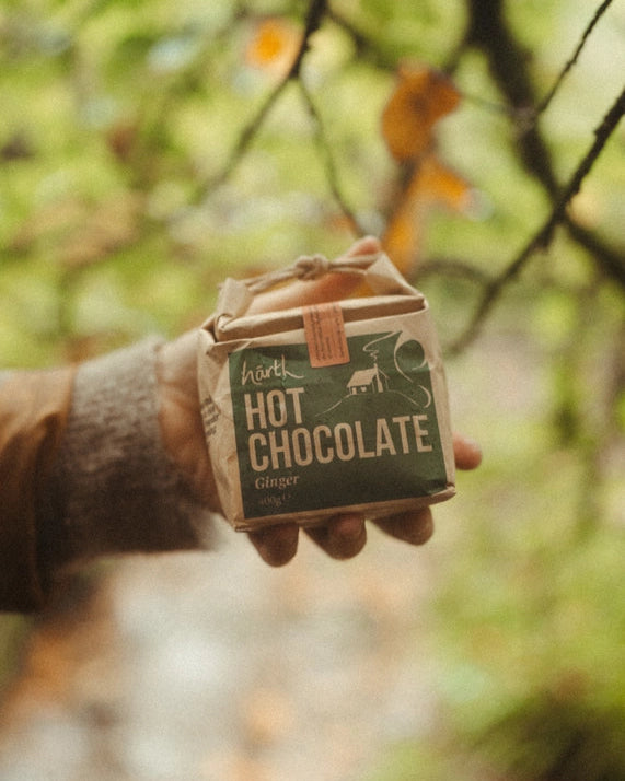 Hand holding Artisan Hot Chocolate – Ginger, a kraft-paper wrapped package with a green label, set against an autumnal outdoor background.