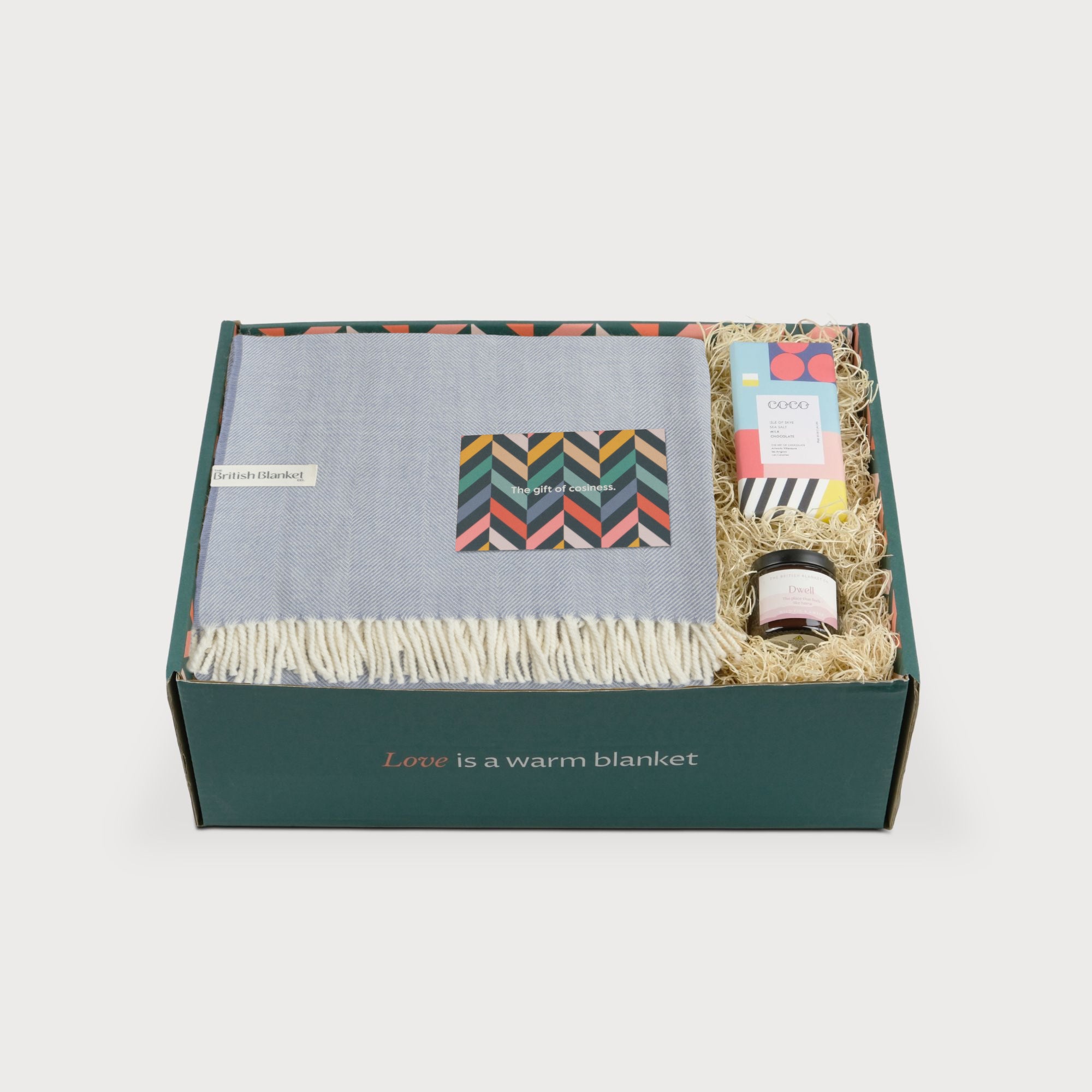 Merino Blanket Gift Box: Smoke Blue with a folded pale-blue throw, geometric card, chocolate bar, and Dwell jar, all in green box.