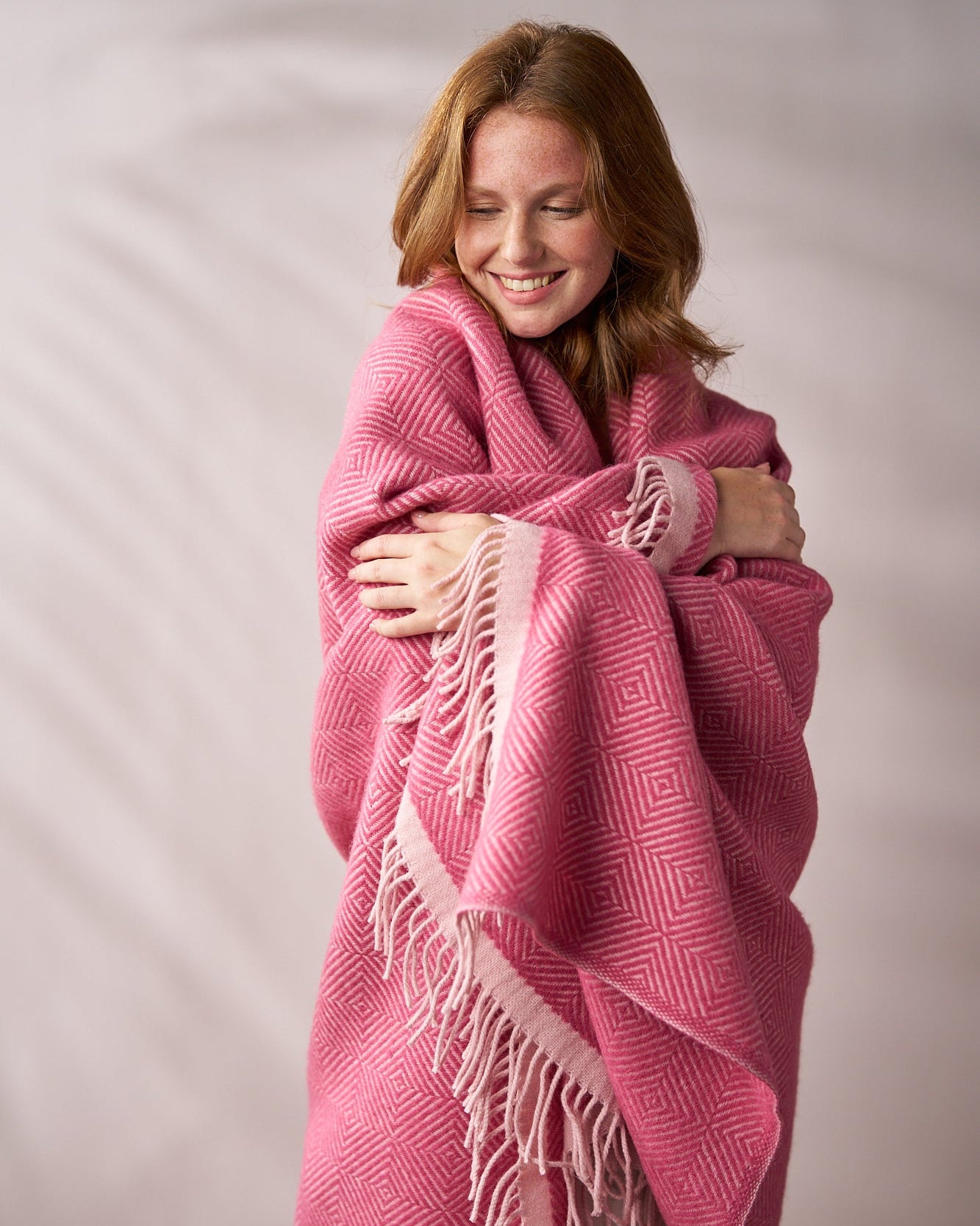 Young woman wrapped in Wildweave Blanket Gift Box: Clover & Briar Rose, hugging it contentedly, showcasing its cozy, fringed design and geometric pattern.