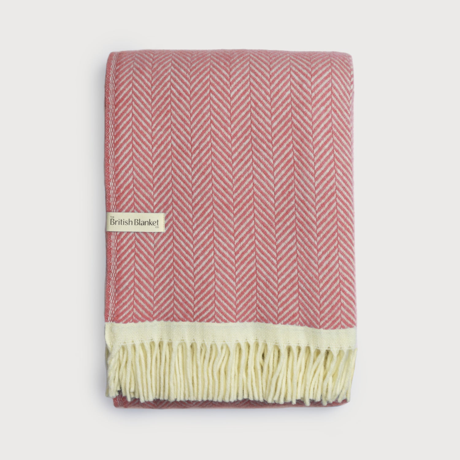 Clifton Herringbone Blanket: Rose, folded in a salmon-pink and cream herringbone pattern, features a cream fringe and a label from The British Blanket Co.
