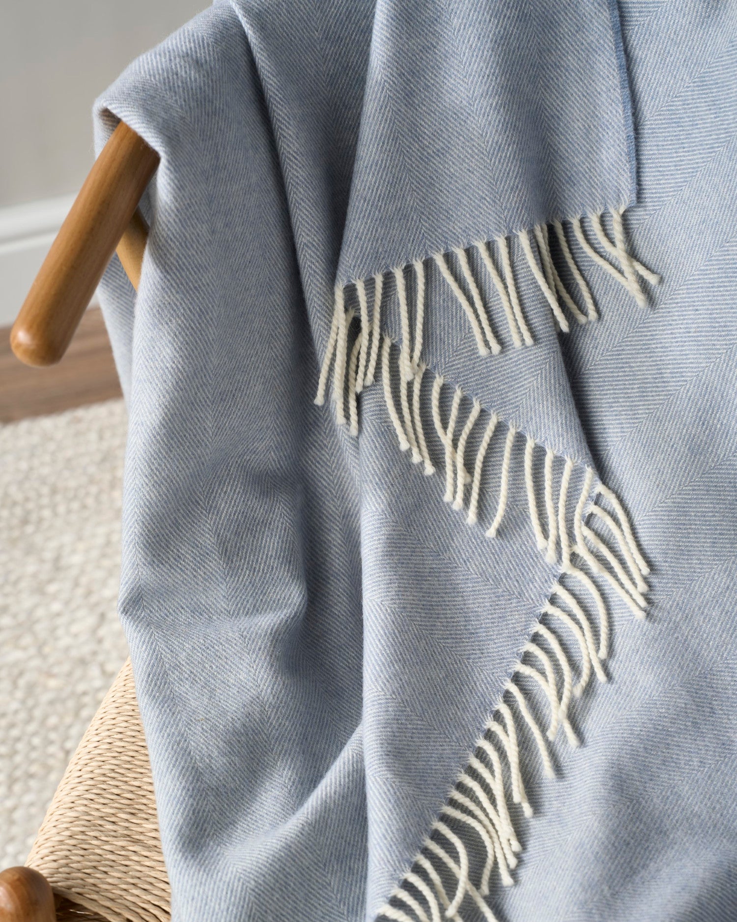 Smoke Blue Merino Blanket Gift Box: A pale-blue herringbone throw with cream fringes on a wooden chair, showcasing luxury for special occasions.