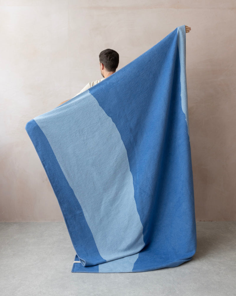 Person holds up Cloud Cotton Blanket: Tide in Cornflower, showcasing its broad, irregular blue bands. Made from recycled cotton, it features a soft, brushed finish.