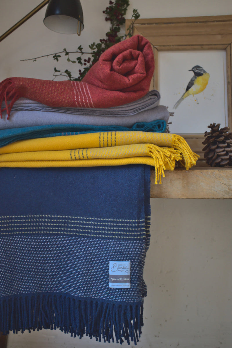 British Birds 'Blue Tit' Merino Lambswool Blanket features vibrant colors, rolled on a wooden shelf with other throws, under a black desk lamp.