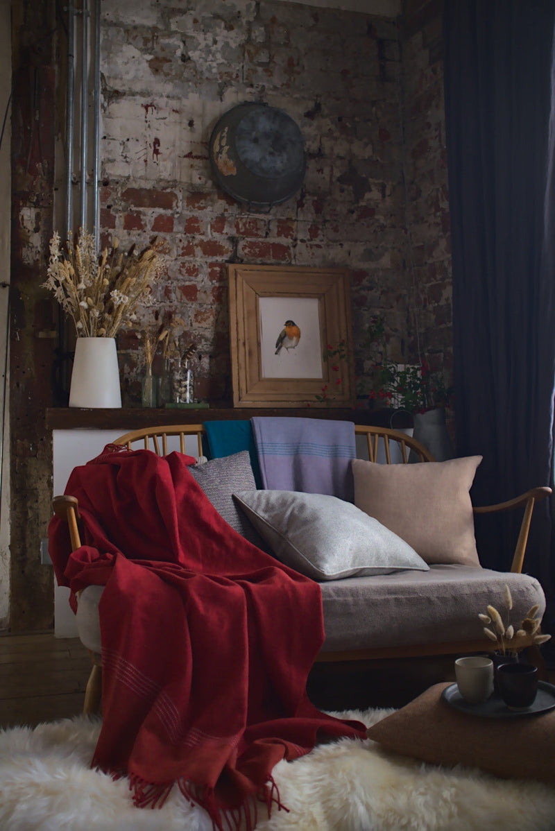 British Birds 'Robin' Merino Lambswool Blanket draped on a wooden settee, beside a sheepskin rug and rustic décor, highlighting cozy, natural vibes.