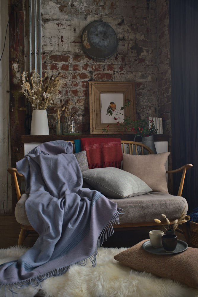 British Birds 'Woodpigeon' Merino Lambswool Blanket drapes a rustic bench; cozy nook features cushions, dried grasses, and a sheepskin rug for a warm, inviting atmosphere.