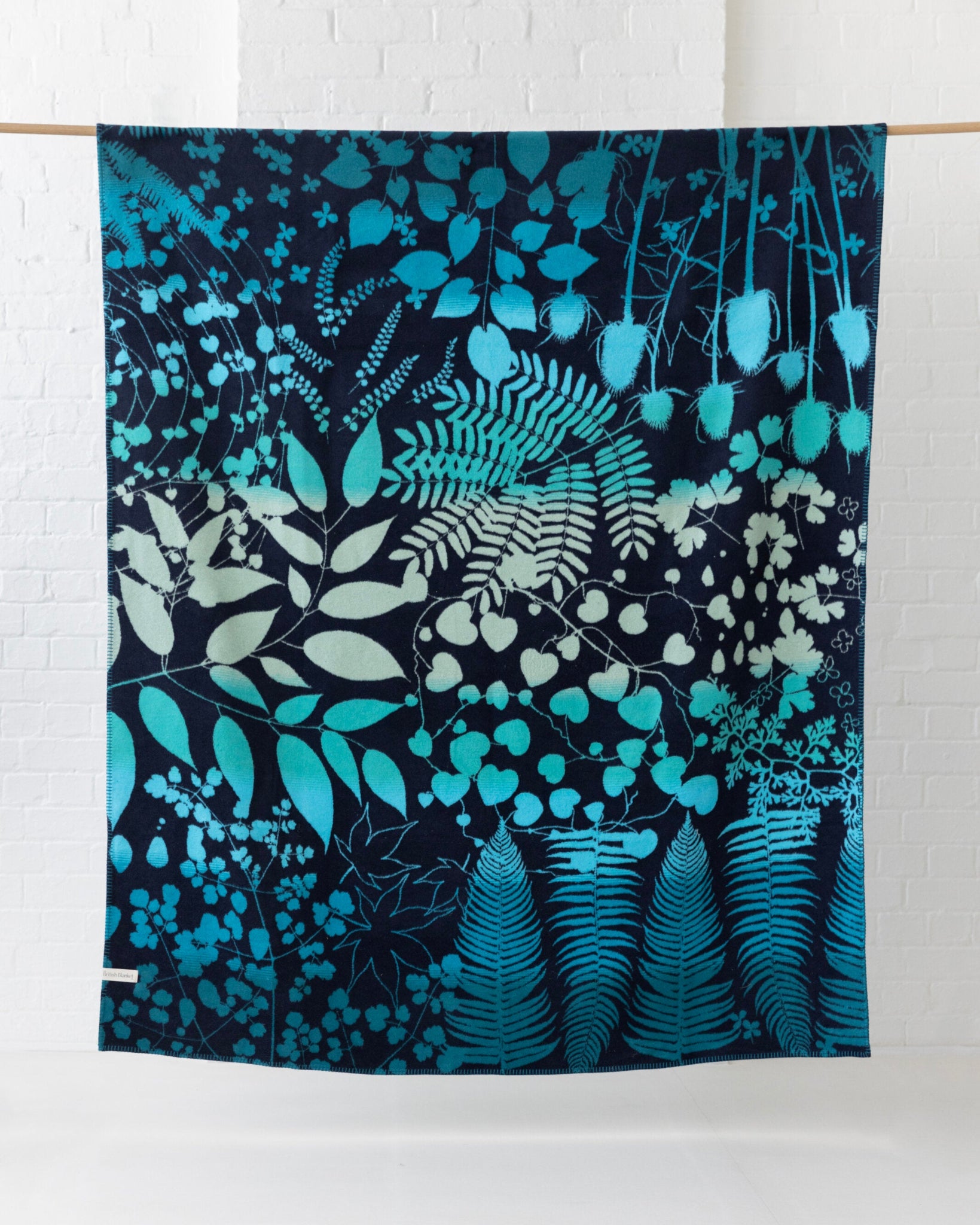 Clarissa Hulse Blanket: Indian Summer in Peacock, showcasing teal and mint botanical silhouettes on navy, hangs elegantly on a wooden rod, suggesting a retail display.