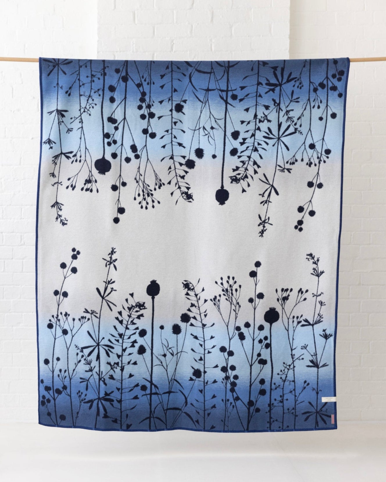 Clarissa Hulse Blanket: Seedheads in Midnight displayed with botanical silhouettes over a gradient fabric, hanging elegantly from a dowel against a brick wall.
