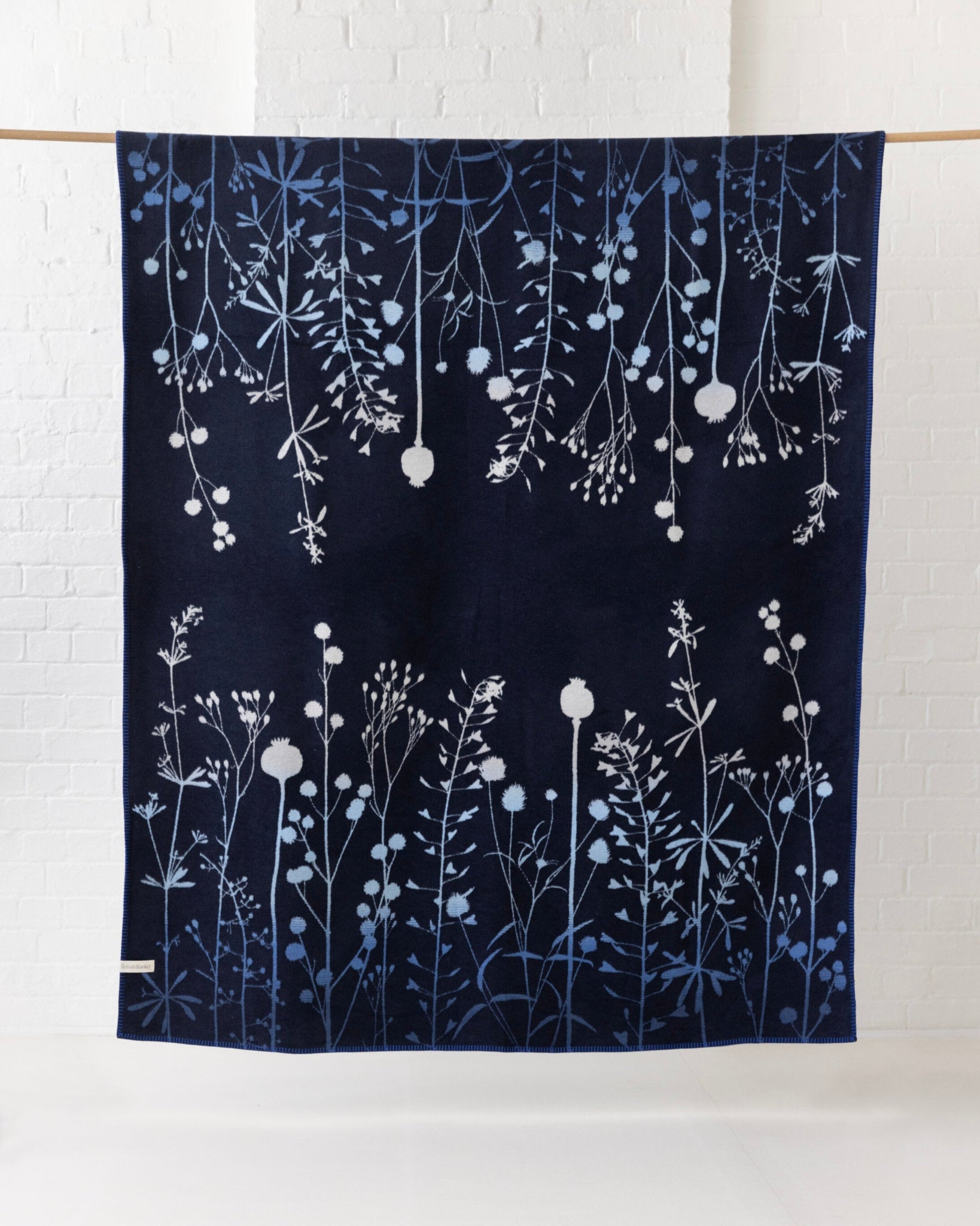 SECOND Clarissa Hulse Blanket: Seedheads in Midnight, featuring a mirrored botanical design with silhouettes of wildflowers and grass stems, displayed on a wooden rod.