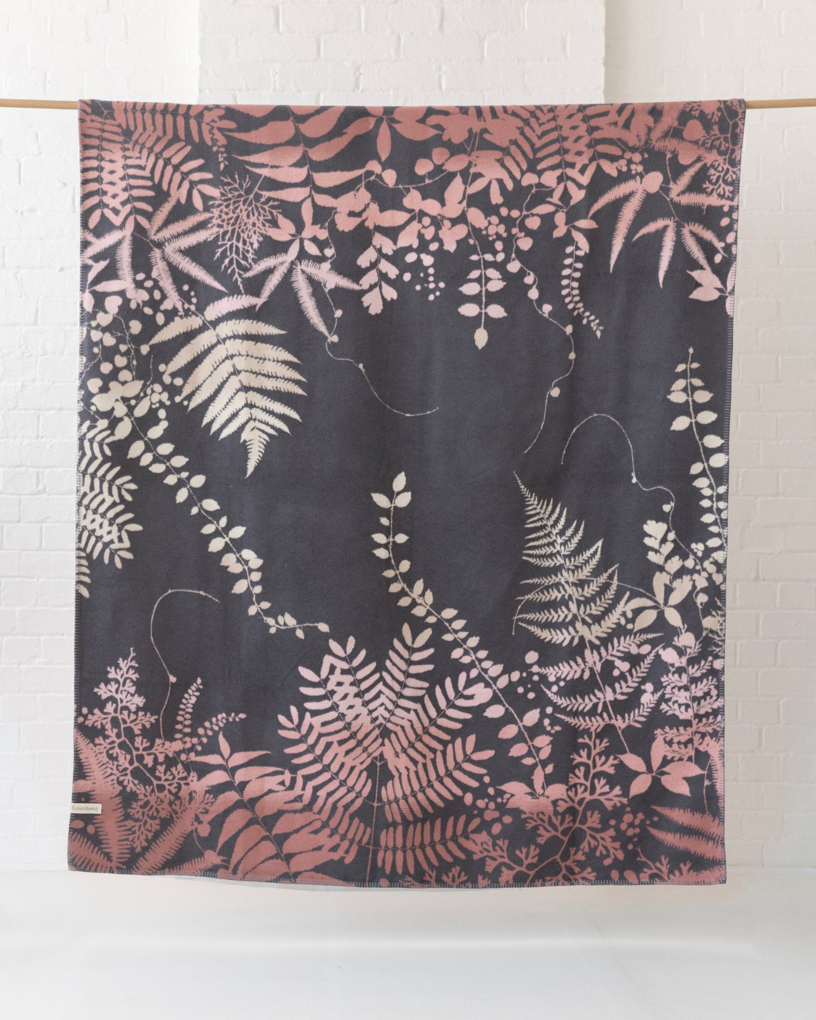 Clarissa Hulse Blanket: Kaleidoscope in Shell features fern motifs in soft pink and cream on a charcoal background, hanging elegantly from a wooden rod.