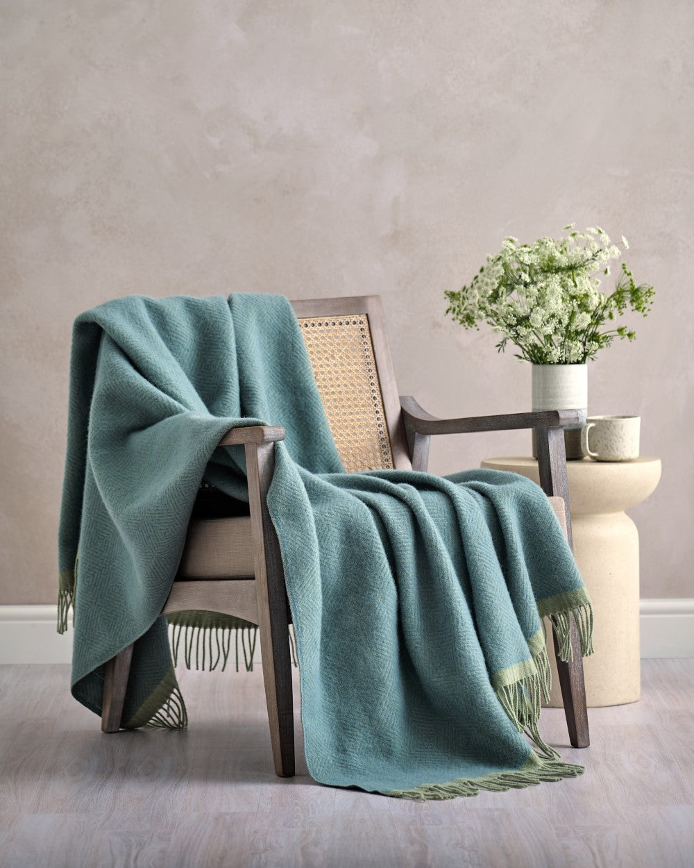 Wooden-framed armchair with a Wildweave Blanket in Sea Holly & Lichen, fringed edges, beside a table with a ceramic mug and vase.