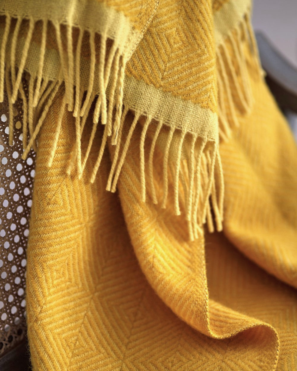 Wildweave Blanket Gift Box: Buttercup & Primrose features a mustard-yellow woven throw with a geometric pattern and fringe, draped over a cane-back chair.