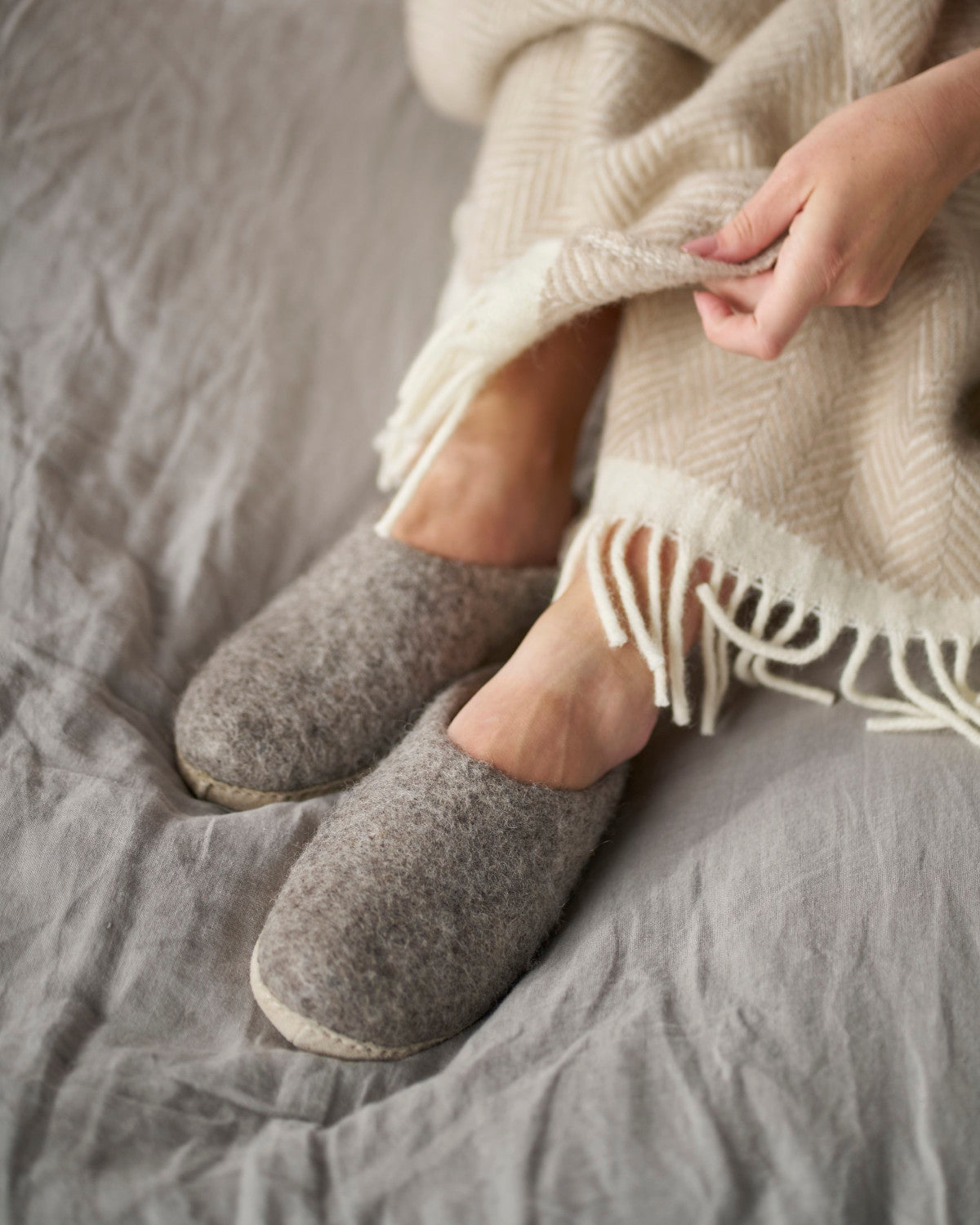 Person wearing Wool Felt Slippers: Natural, wrapped in a blanket, highlighting the slippers' cozy fit and soft suede sole, perfect for indoor use.