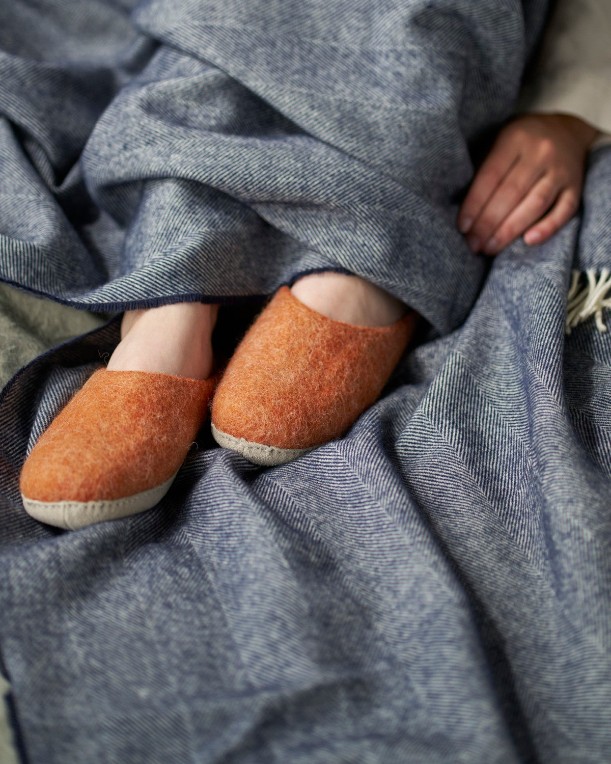 Person's feet wearing Wool Felt Slippers: Clay under a blanket, highlighting the cosy and cushioned design with a soft suede sole, perfect for indoor use.