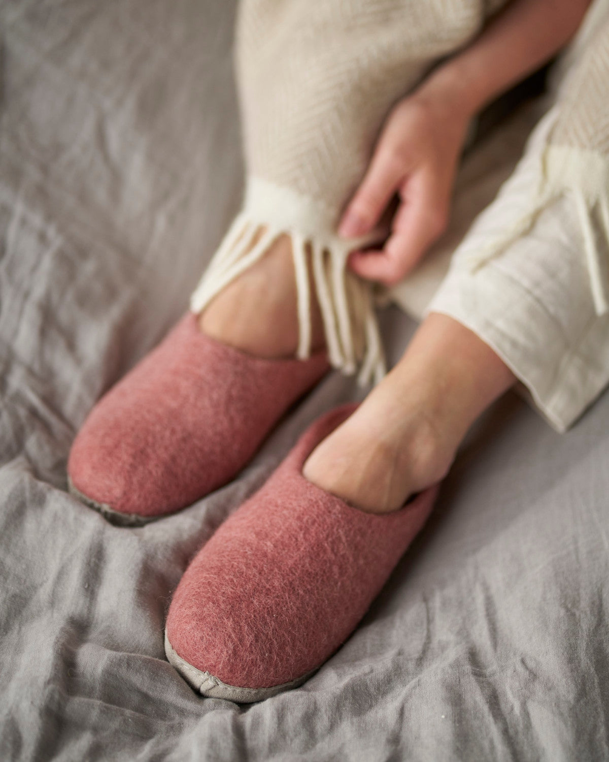 Person wearing Wool Felt Slippers: Rose, with white leggings, holding a blanket. Slippers feature a soft suede sole, ideal for indoor comfort.