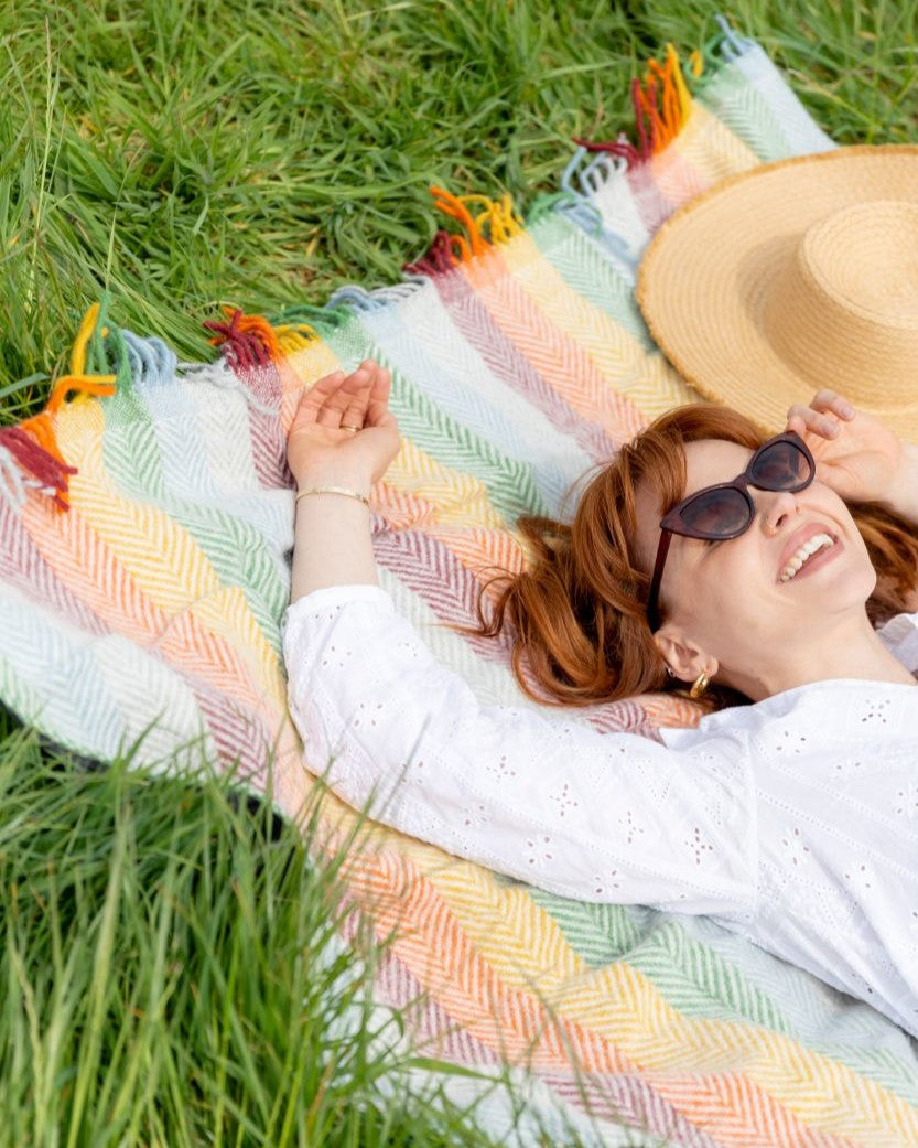 Woman relaxing on a Waterproof Picnic Blanket: Bright, featuring pastel herringbone stripes and fringes, on grass; includes a straw hat and leather straps.