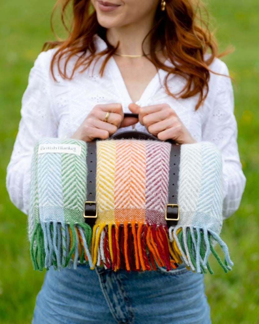 Woman holding Waterproof Picnic Blanket: Bright, featuring a herringbone wool design in rainbow stripes, leather straps, and a British Blanket label, in a grassy field.