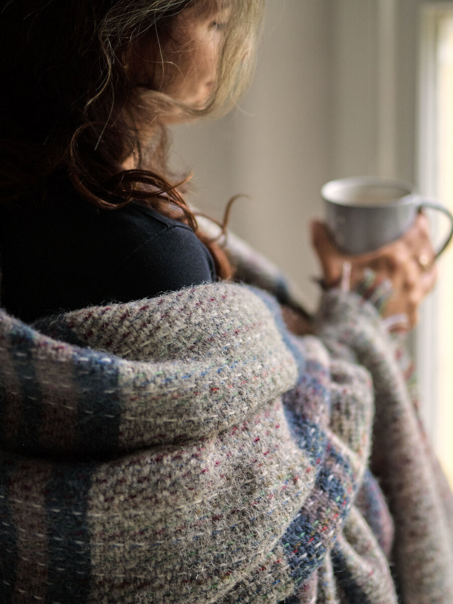 A person wrapped in a Random recycled wool blanket (C), holding a ceramic mug, suggests warmth and comfort while standing indoors.