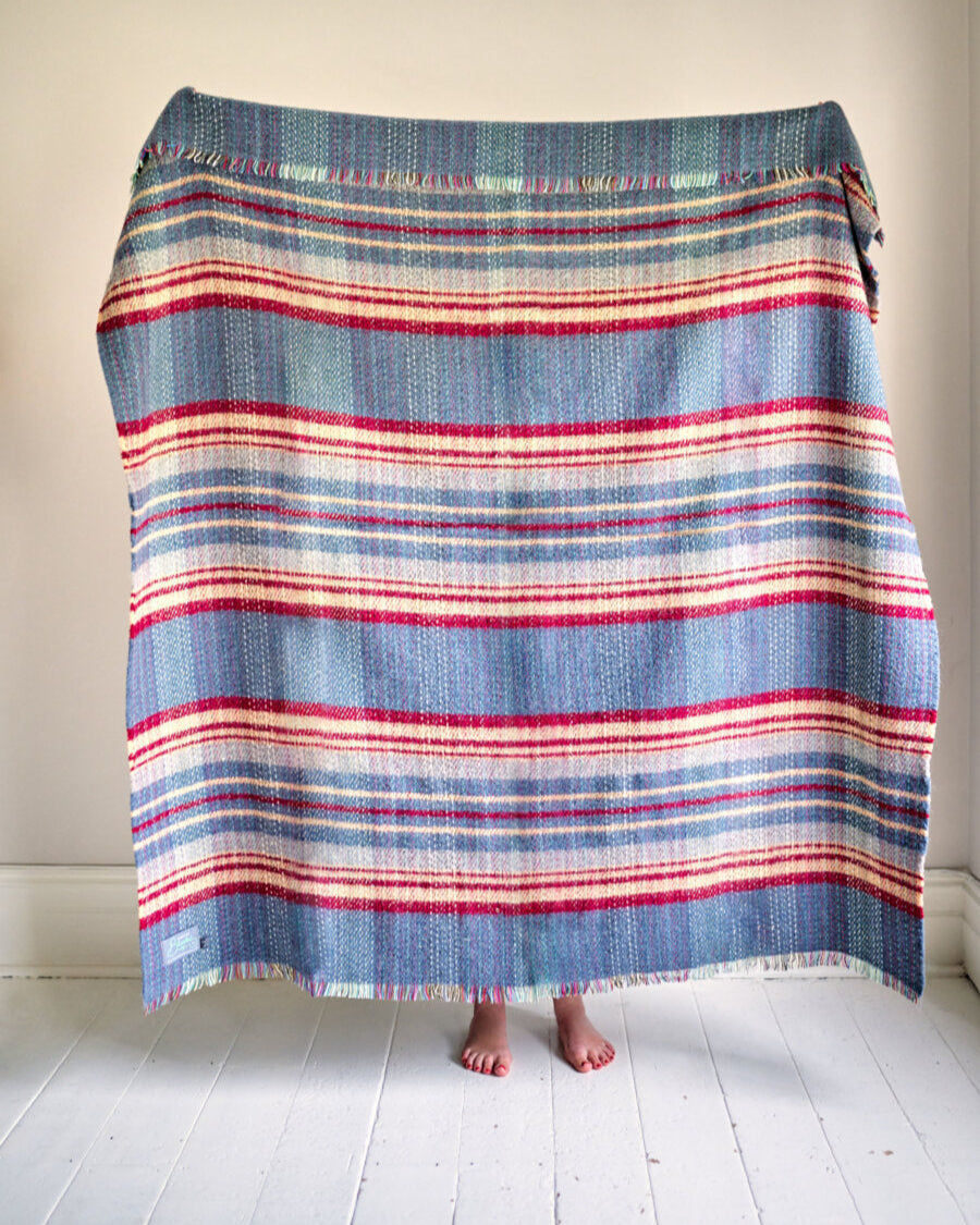 Cosy Crew Reward: 3 For 2 Random Recycled (Large) blanket, hand-woven with stripes, fringed edges, held up showing unique design, bare feet visible below.