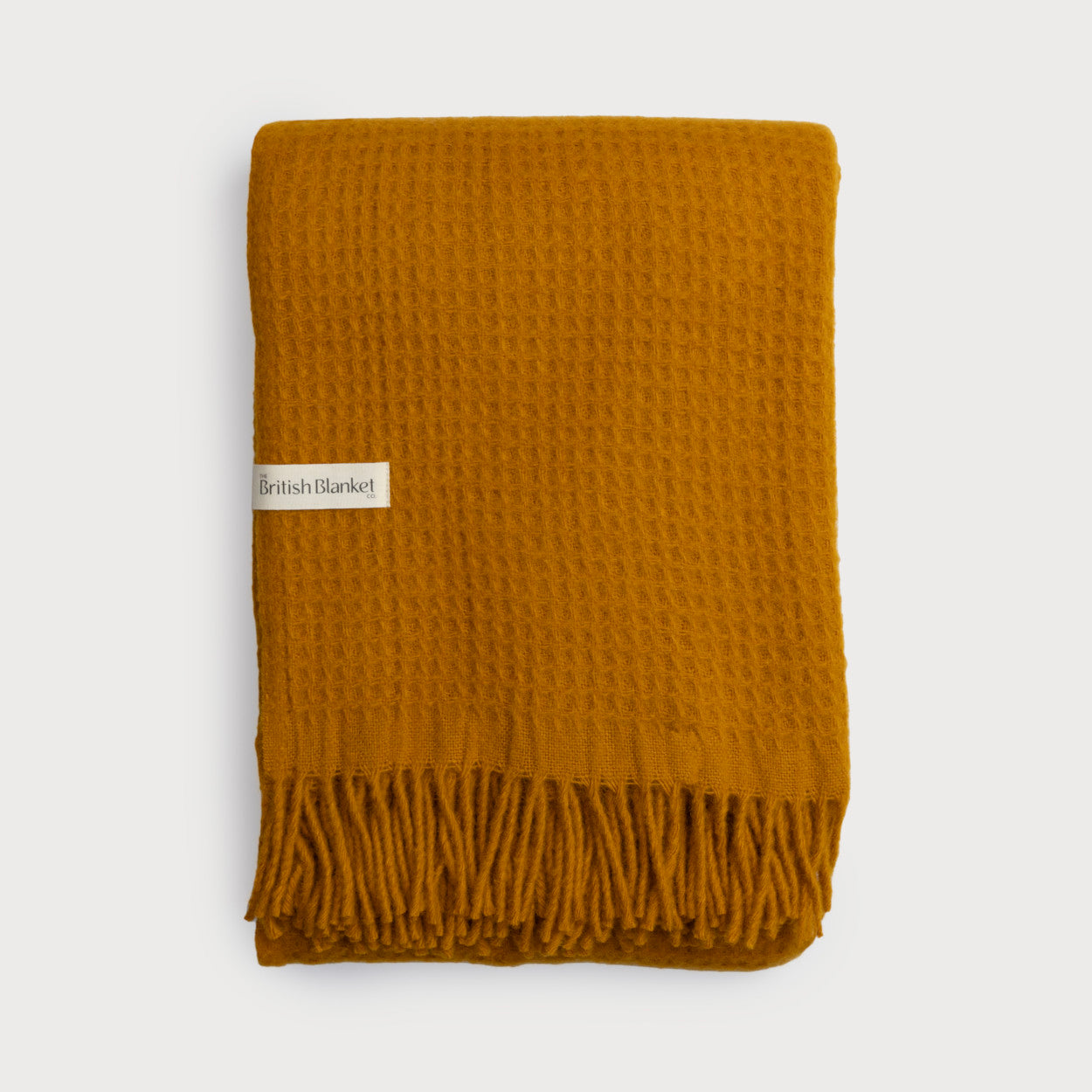 Folded Bright Friday Blanket Bundle: Ashton with waffle-weave texture, mustard-yellow wool, and knotted fringe. Features British Blanket Co. label on the side.
