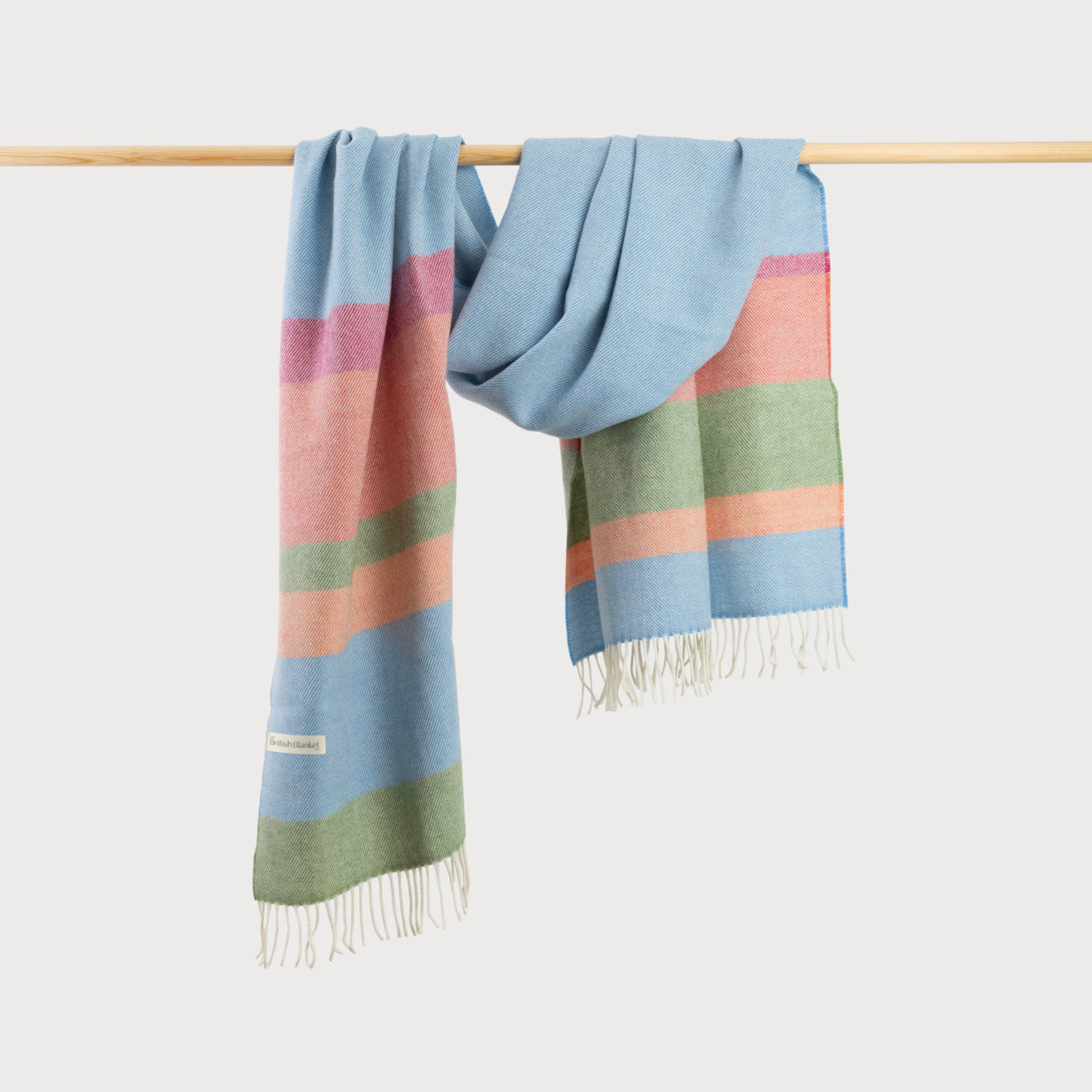 Oversized soft merino lambswool blanket scarf in blue, green, pink and white striped pattern, hanging on a rod.