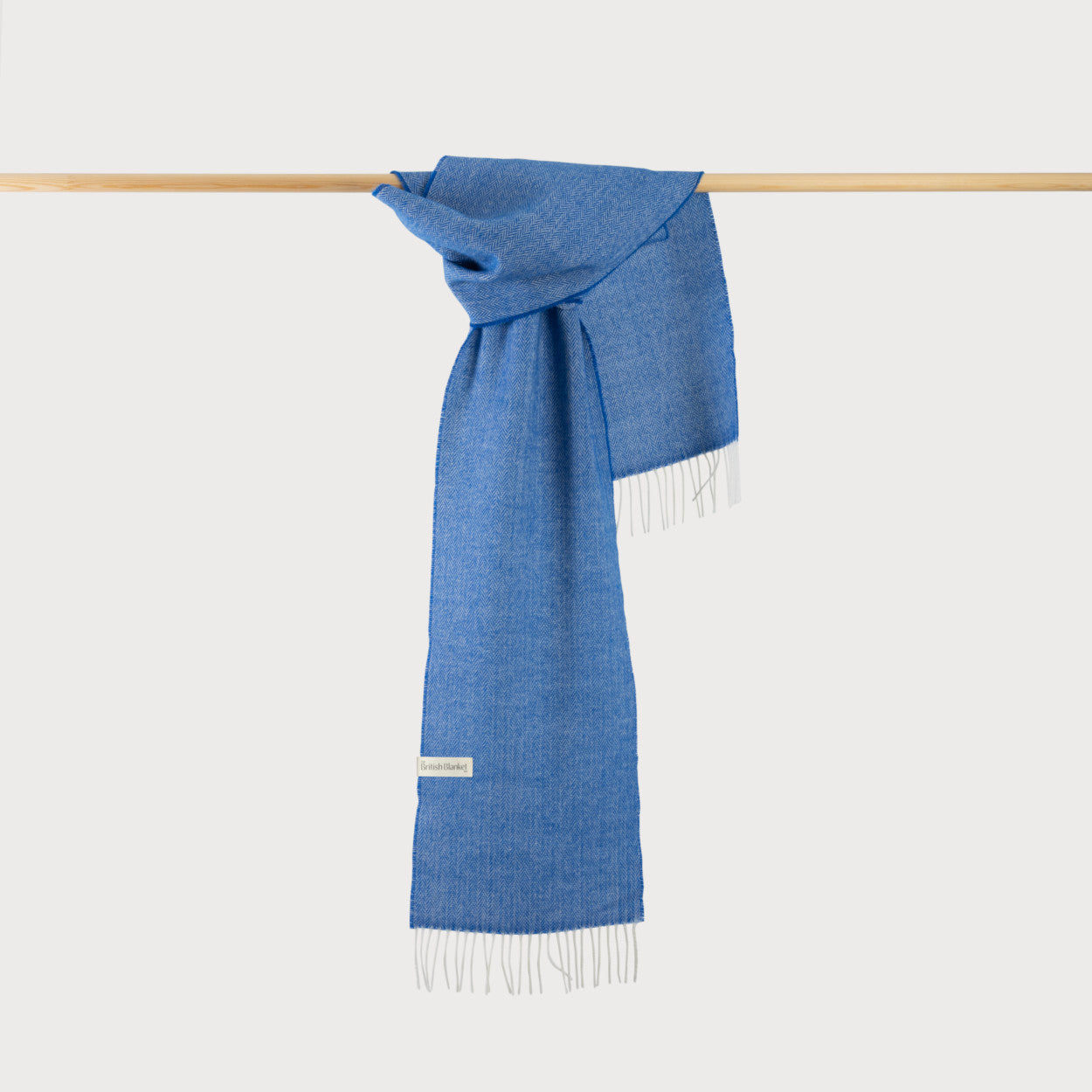 Herringbone Scarf Gift Box: Cobalt Herringbone features a light-blue woven scarf with white fringe, elegantly draped over a wooden rod, showcasing its merino wool texture.