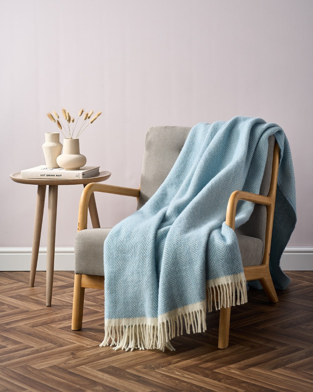 A cozy reading nook featuring a gray armchair with a pale-blue herringbone-patterned Clifton Blanket Hamper: Harbourside draped over it, beside a wooden side table with vases.