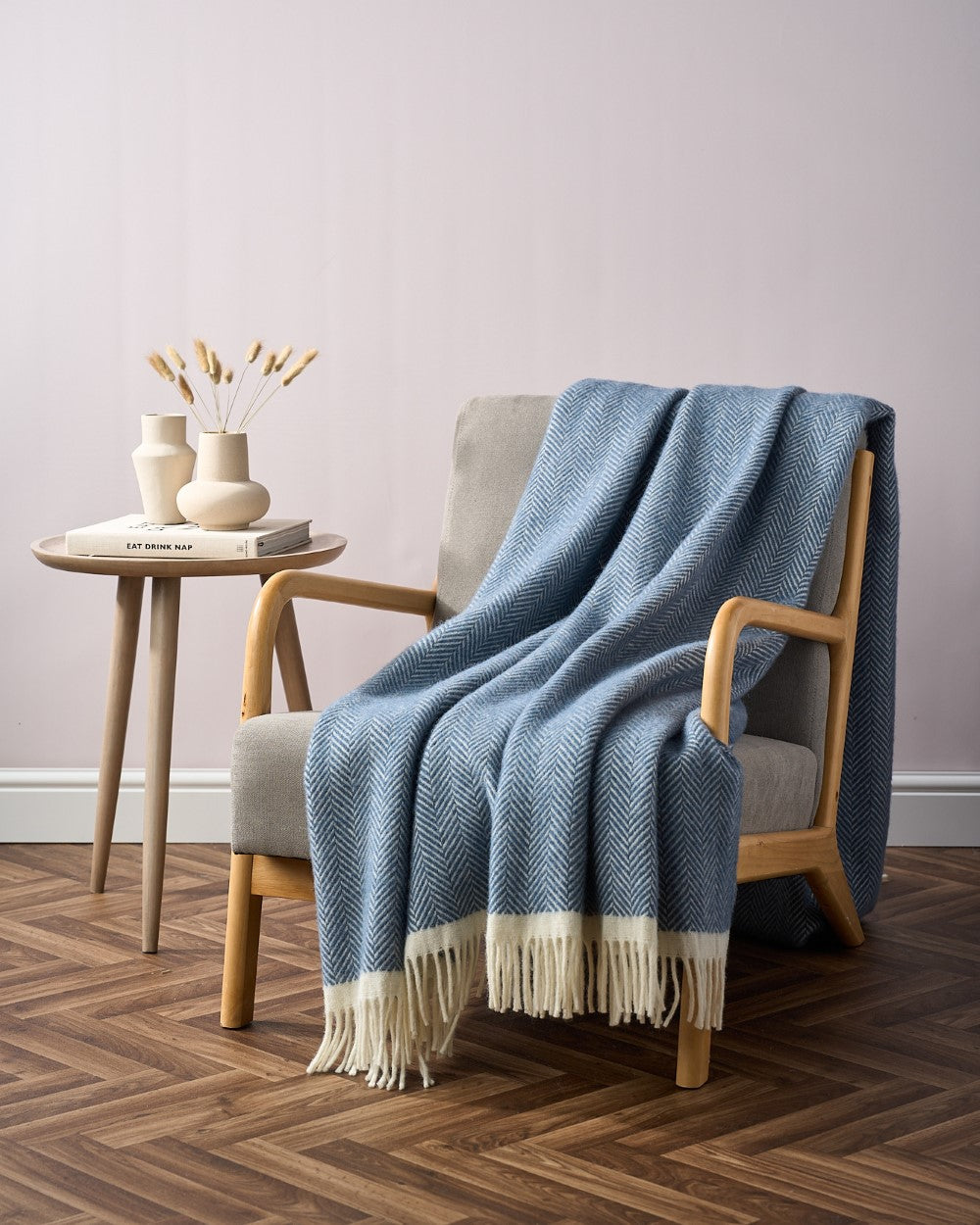 A cozy living space featuring the Clifton Blanket Hamper: Steamship, a blue herringbone-patterned throw with fringed edges draped over a light-wood armchair.