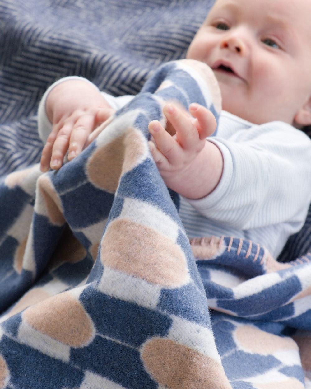 Baby with Cloud Cotton Baby Blanket Gift Box: Boatyard Shell, featuring a navy, tan, and off-white geometric pattern, lying on a textured blue blanket.