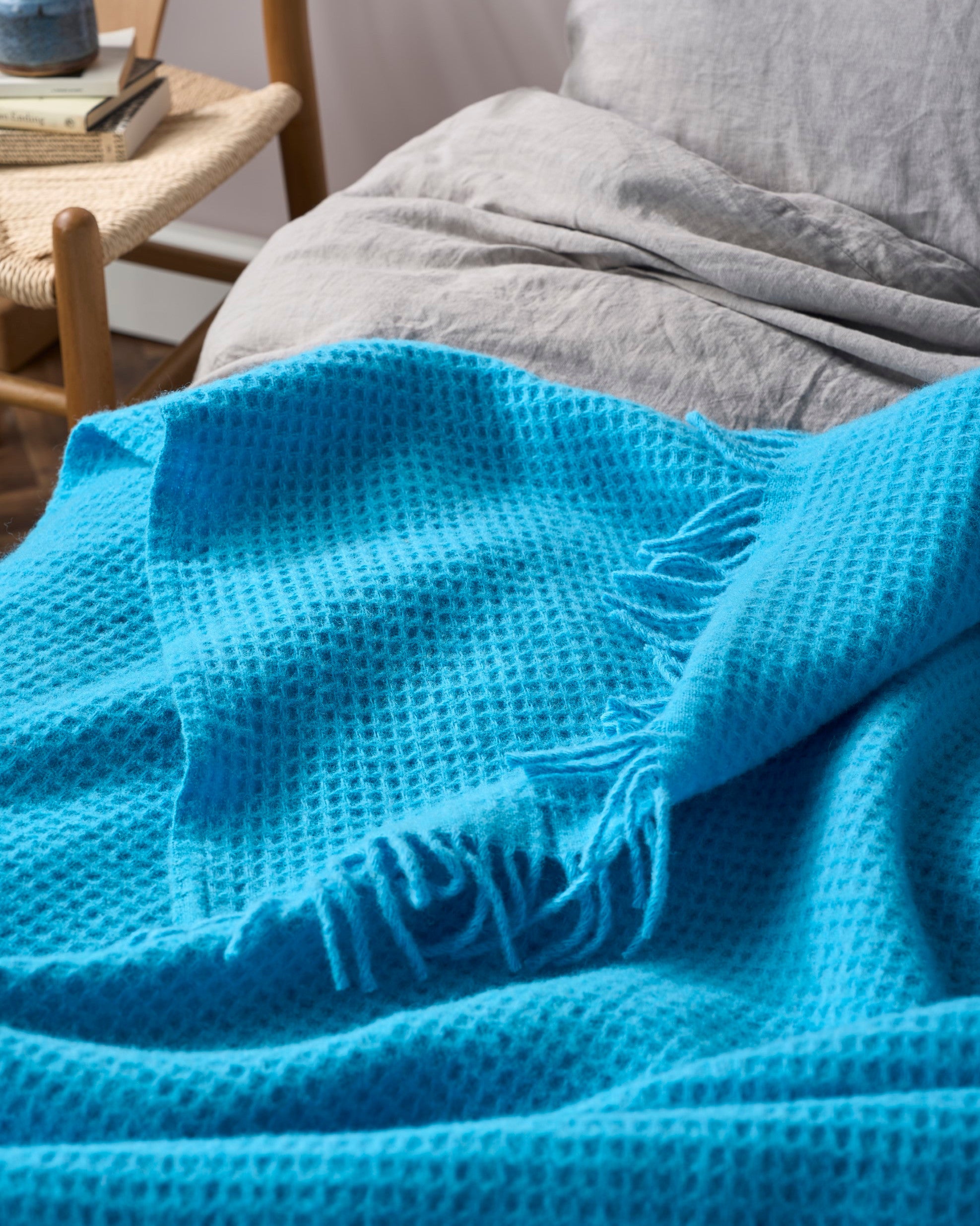 Bright Friday Blanket Bundle: Lido on a bed with light gray sheets, featuring a turquoise waffle-weave throw with fringe. Nearby, a chair holds books and a ceramic pot.