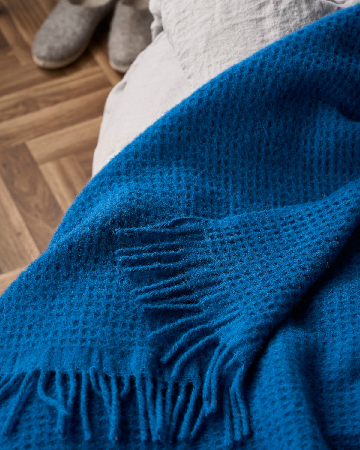 Bright Friday Blanket Bundle: Curious features a textured woven throw with a waffle pattern and fringed edges, draped over a bed, complimented by nearby soft slippers.