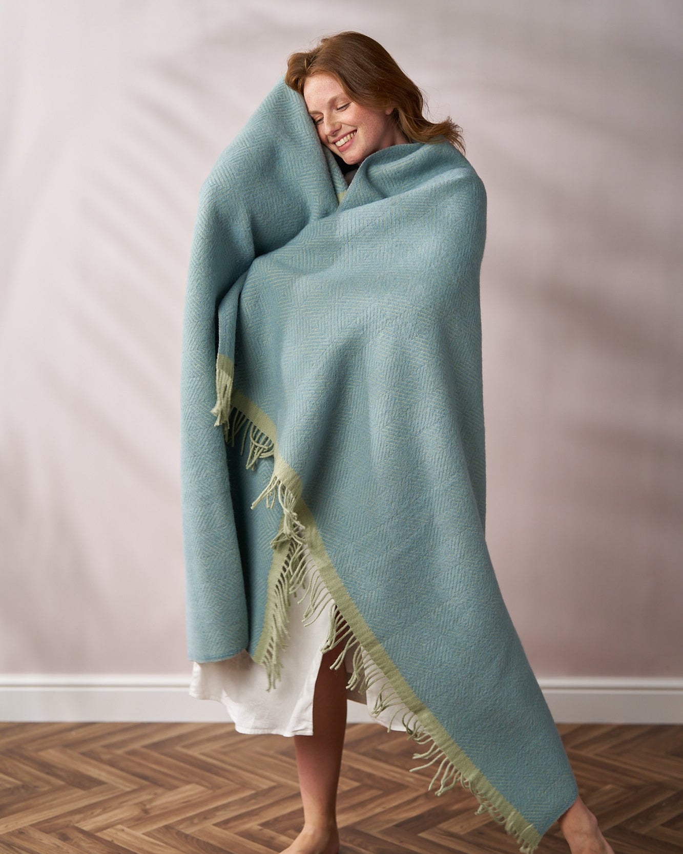 Person wrapped in a light-blue Wildweave Blanket Gift Box: Sea Holly & Lichen, standing barefoot on wood floor, smiling and holding the blanket close.