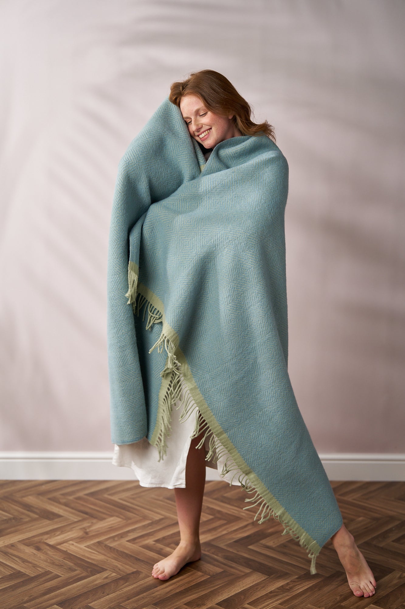 Person wrapped in a light-blue Wildweave Blanket Gift Box: Sea Holly & Lichen, standing barefoot on wood floor, smiling and holding the blanket close.