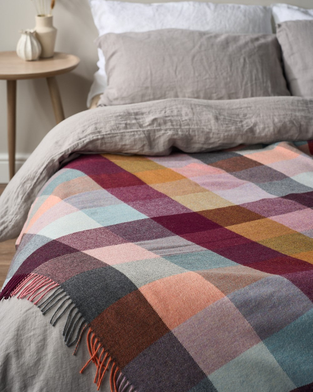 Merino Check Blanket: Burnished, featuring a checked pattern and multicolored tassels, draped on a bed with pillows and a blanket.