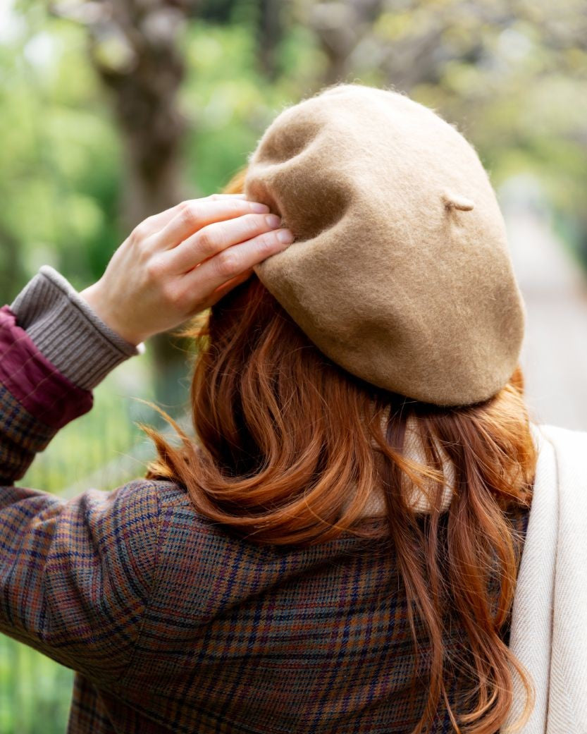A woman adjusts the Camel Brown Wool Beret Hat, showcasing its soft, structured felt in an outdoor setting, complementing her plaid coat.