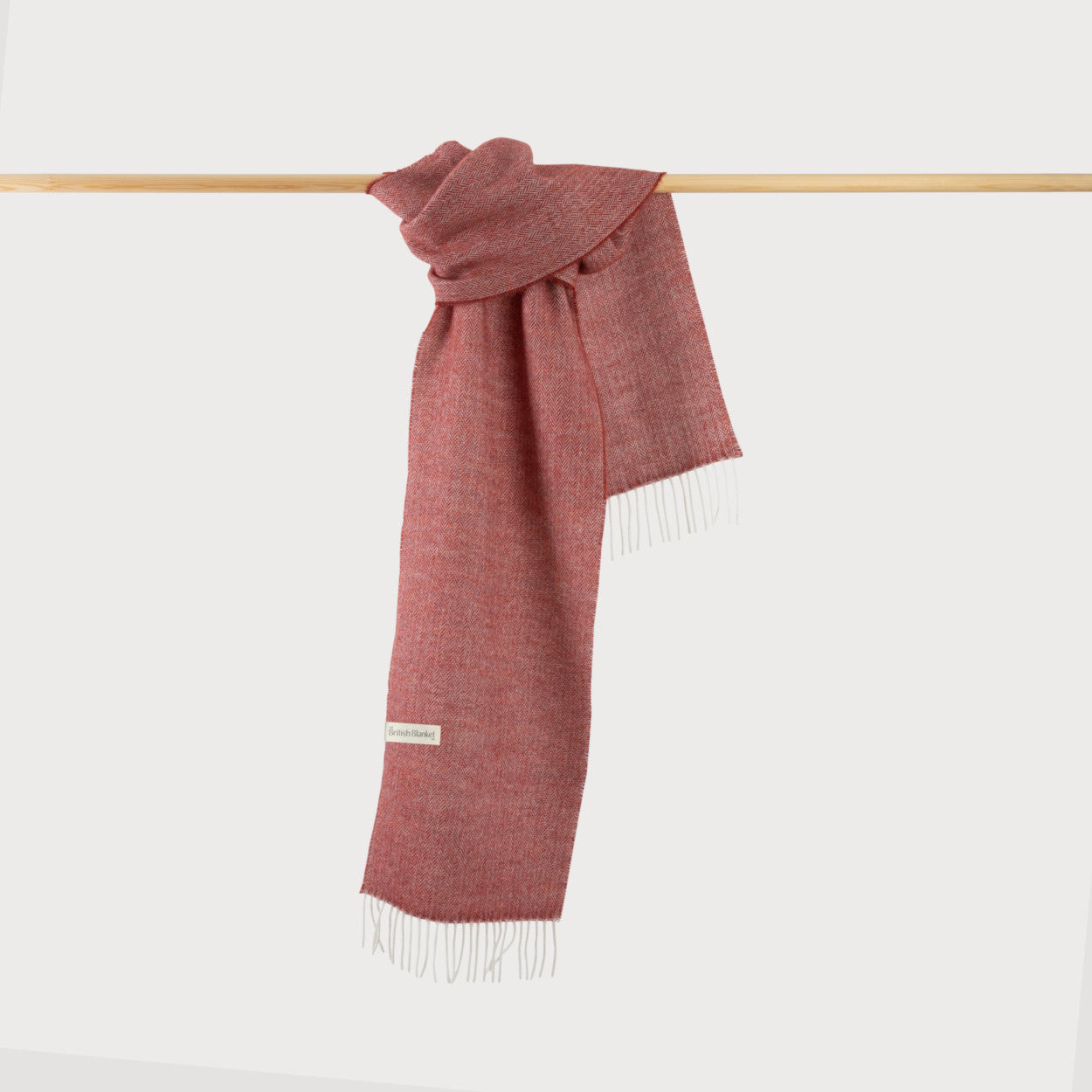 Herringbone Scarf: Chilli, displayed on a wooden pole, showcasing its fine herringbone weave and white fringe, made from soft merino lambswool.