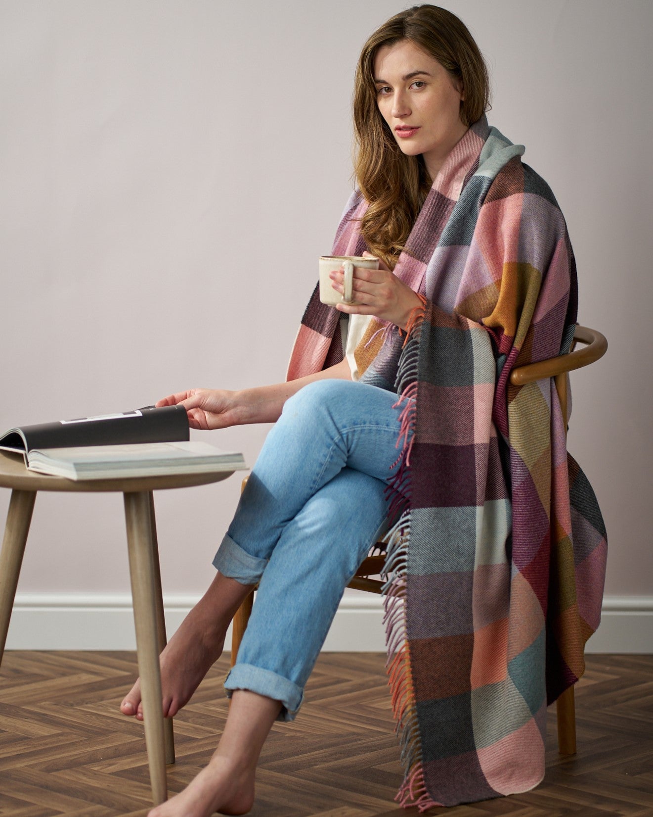 Woman sitting in a chair with a Merino Check Blanket: Burnished, holding a cup and a book, showcasing its soft, luxurious texture.