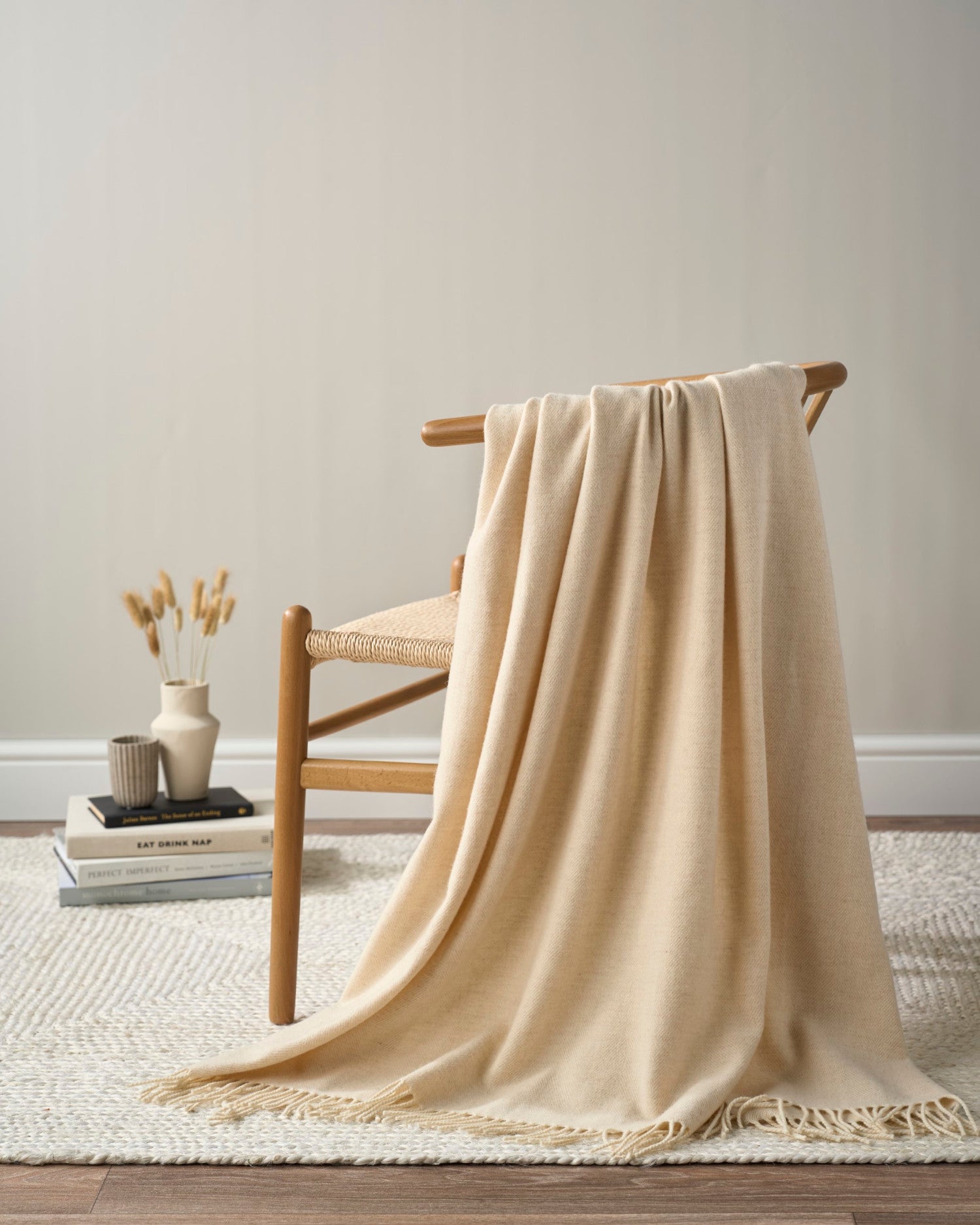 Merino Blanket Gift Box: Cream features a woven chair with a beige throw, books, vases, and dried grasses, embodying warmth and natural elegance.