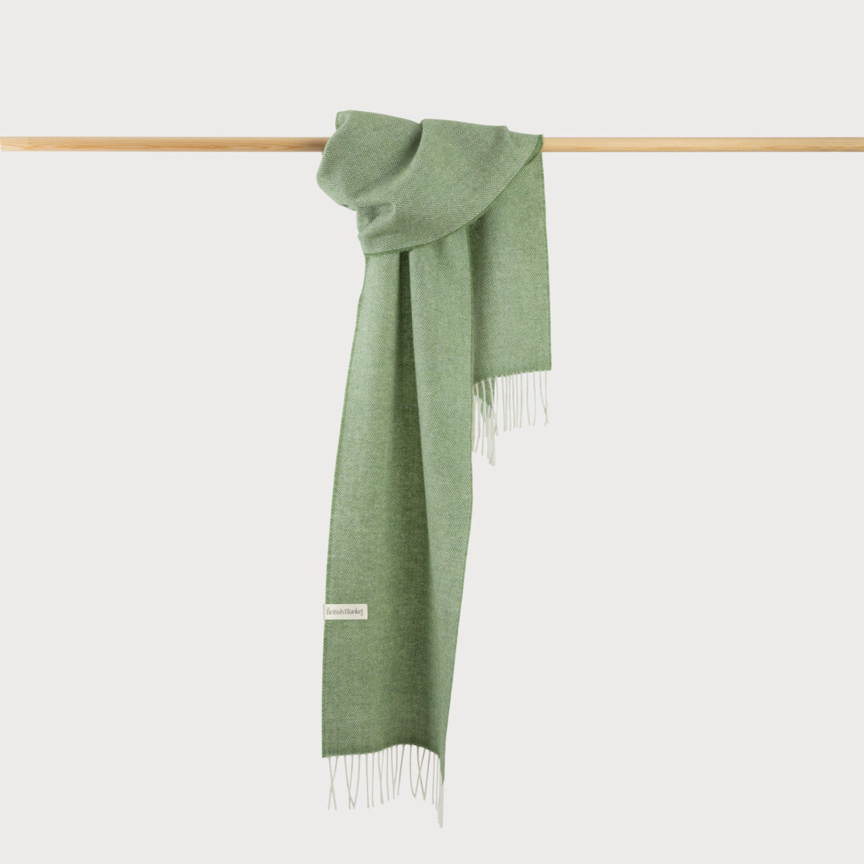Scarf Gift Box: Apple Herringbone features a muted green herringbone scarf with fringe, woven label, draped on a wooden rod, showcasing its soft texture.