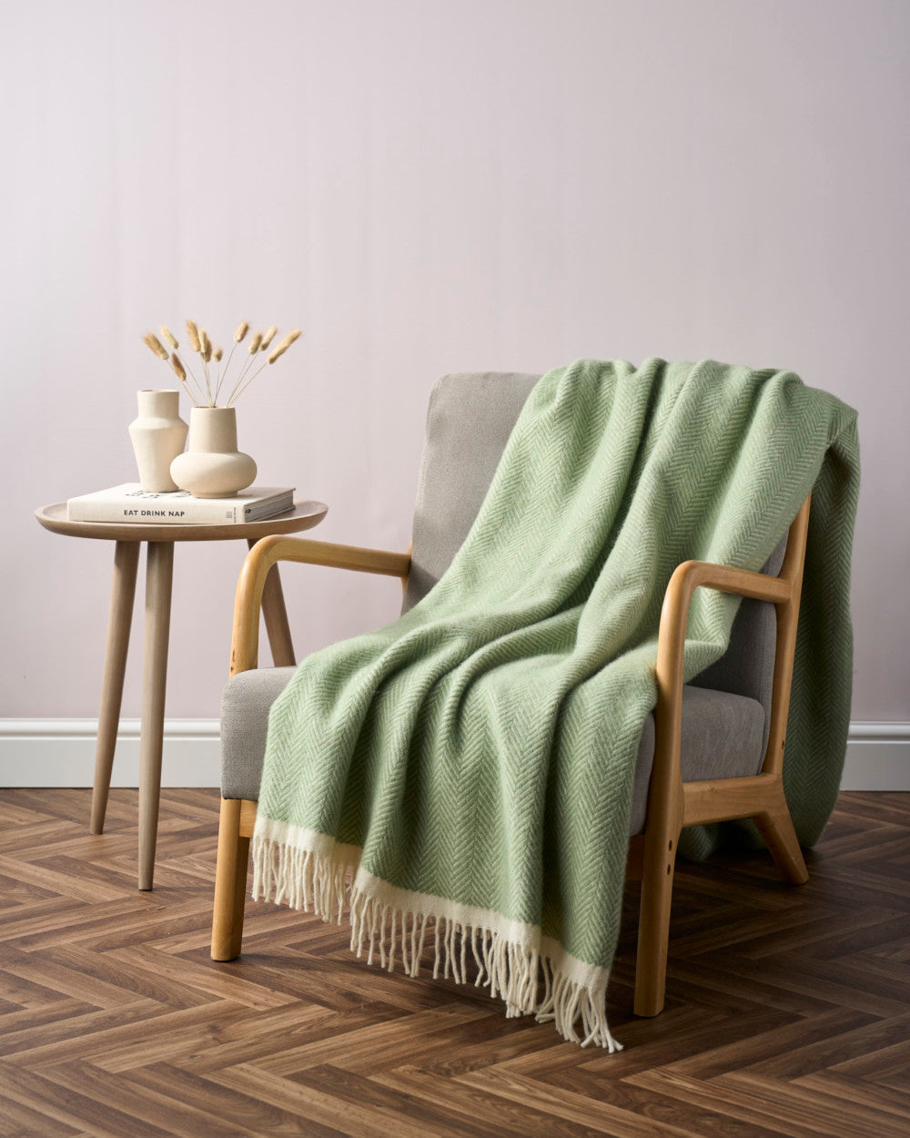 Clifton Herringbone Blanket: Birdcage draped over a grey armchair, showcasing its pale-green herringbone pattern and cream fringes, beside a wooden table with ceramic vases.