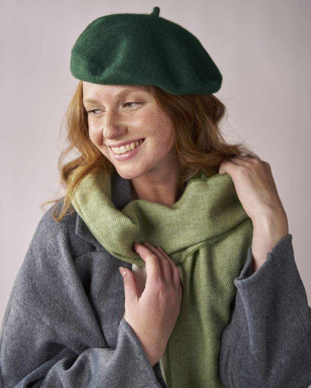 Young woman in a soft green beret titled slightly, made of thick, durable wool, paired with a matching herringbone scarf and gray woolen coat.